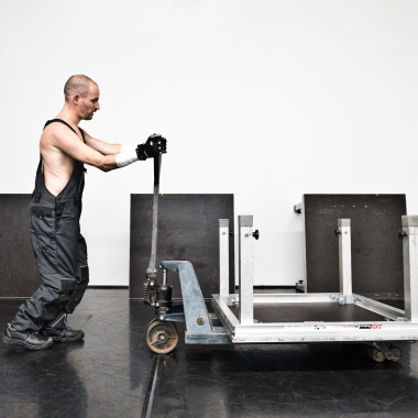 A scene from Work Body: It shows Michael Turinsky on stage. He is wearing dungarees and pushing a trolley in front of him.