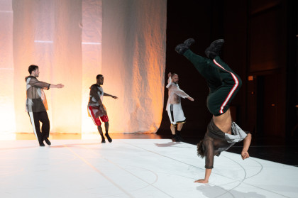 A scene from ‘Il Cimento dell'Armonia e dell'Inventione’: Four dancers on stage. They are all wearing loose, sporty clothing. One dancer performs a handstand in the foreground, the other three stand next to each other in the background, each dancer performs a rotation.