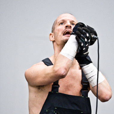 A scene from Work Body: It shows Michael Turinsky with a microphone, which he is holding tightly with his hands. He is wearing dungarees and is looking up.