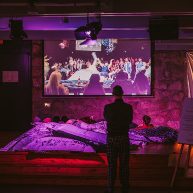 The festival centre in the foyer of Ballhof Eins from the inside. It is bathed in purple light. In the centre of the picture is a standing person from behind, looking at a large screen showing an event. In front of the person is a stage with several white beanbags in which some people are sitting.