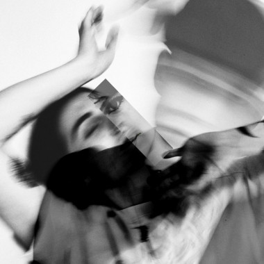 A black and white photo from the work FOR REAL#2 Trazos: It shows a dancing person holding their arms in the air and closing their eyes. Half of another face has been collaged onto the person's face. The image is slightly blurred. The shadow of another person is visible on the picture.