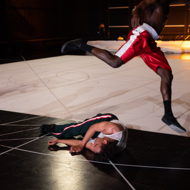 A scene from the play ‘Il Cimento dell'Armonia e dell'Inventione’: Two dancers, dressed in sporty clothes, on stage. One dancer is lying on the black and white stage floor, on which various lines can be seen. The other dancer jumps into the air and is wearing red sports trousers. His head is not seen in the picture. In the background, the stage is illuminated with warm light. 