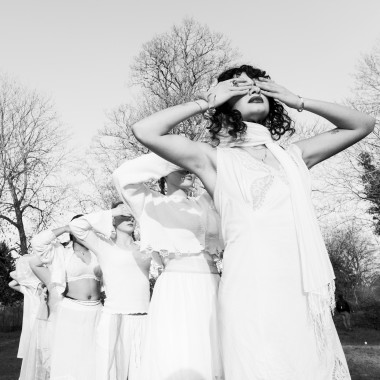 A photo in black and white of several people standing one behind the other outdoors, they each cover their eyes.