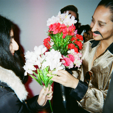 A scene from Keeping up with the Iranians: In the right half of the picture, it shows a performer from the play handing a bouquet of flowers to the person opposite to them. Both are smiling. 