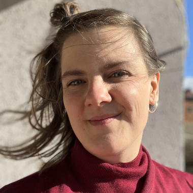 A portrait of workshop leader Dr Anna Wieczorek. Anna is wearing a red turtleneck jumper and has long brown hair. She smiles into the camera.