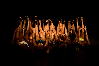 A photo that shows a scene from the play VAGABUNDUS by Idio Chichava. It shows several dancers on a dark stage. They are lying on their backs next to and behind each other, stretching their legs in the air at the same time. They are all wearing shorts.