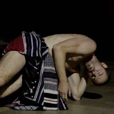 A scene from the video work TRANSEPT (2023) by Boris Charmatz & César Vayssié. It shows Boris Charmatz on the floor with a colourful patterned scarf tied around his hips. His eyes are closed.