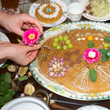 Ein großer Teller Halva. Die Süßspeise ist aufwendig verziert mit Früchten, Blumen und Nüssen. Daneben kleinere Teller mit anderen Speisen.In der linken Bildhälfte zwei Hände, die eine Blume halten.