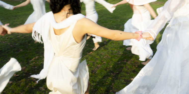 Several people in white clothing stand hand in hand in a circle on a green area.