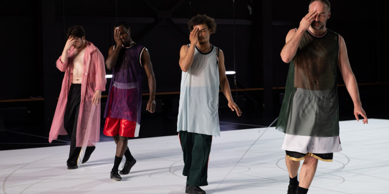 Four dancers stand next to each other on the stage. They each cover their faces with one hand. They are all wearing loose, sporty clothing.