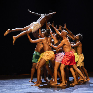 A scene from VAGABUNDUS: It shows thirteen dancers on stage wearing colourful shorts. They stand together on a surface and throw one person from the group into the air. The person in the air extends their arms.