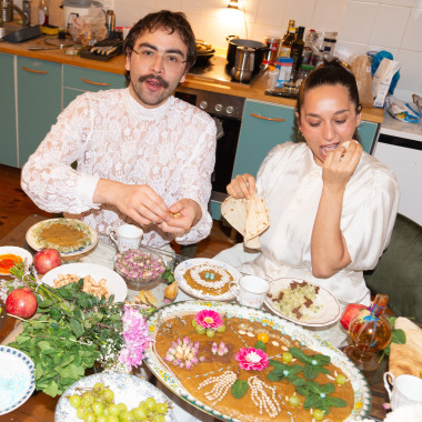 Ein Foto, das Afrang Nordlöf Malekian (links) und Sepideh Khodarahmi (rechts) zeigt. Sie sitzen ganz in weiß gekleidet in einer Küche. Auf dem Tisch vor ihnen sind viele verschiedene Speisen zu sehen, darunter eine mit Blumen, Früchten und Nüssen verzierte Halva. Afrang blickt in die Kamera, Sepideh beißt in ein Stück Brot.