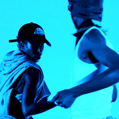 A scene from 1GUH WATCH: Two dancers on the stage, which is bathed in blue light. They are wearing loose, sporty clothing and dancing. 