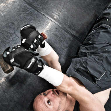 A scene from the play Work Body: The photo shows Michael Turinsky from above. He is wearing dark dungarees and is lying on his back on the floor. He is looking at a hammer that he is holding in his hands. 