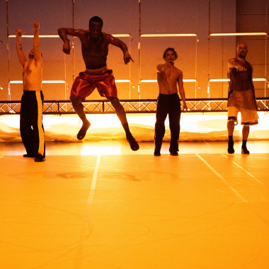 Four dancers stand next to each other on the stage.  They are all wearing loose, sporty clothes. The second dancer from the left leaps into the air. The stage is bathed in warm yellow light.