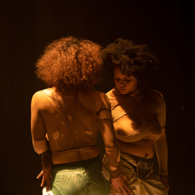 Photo from the production LOUNGE: Two women appear to be walking close to each other. They touch each other lightly with their hands on their hips and thighs. They are wearing cut-up clothes with large holes in them and do not look at each other. The scene appears very intimate and is bathed in dim yellowish light.