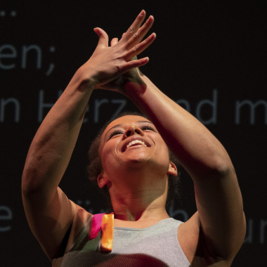 Naomi Sanfo-Ansorge beim Applaudieren. Naomi trägt ein graues Tank-Top und lächelt. Ihre Hände hält sie beim Applaudieren hoch. Hinter ist eine schwarze Wand, auf der Wörter in blauer Schrift zu erkennen sind.  