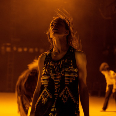 A performer from the production “TARAB”. The person is standing on a stage bathed in orange light with her eyes closed and her mouth slightly open. She throws her head back slightly, her long hair in motion. The performer is wearing a sleeveless dark top with decorative curtain hooks. Two other performers can be seen moving in the background.