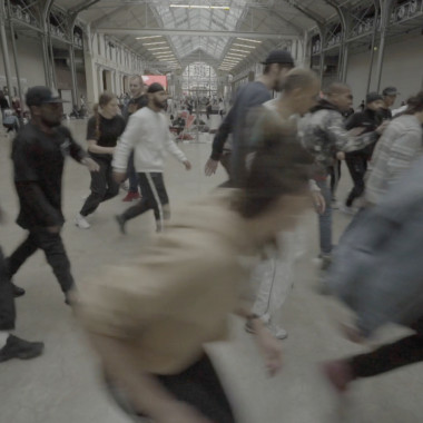 A high room that looks like an empty factory hall. A group of about 15 people move in a crowd from left to right through the picture, they seem to be running. They are all wearing different clothes and trainers. One person on the right of the picture appears to be breakdancing, the eyes of the other people are on the person.