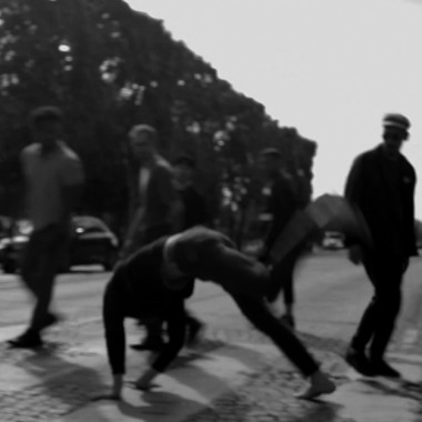 Black and white photo of a situation on a street with cobblestones, trees and cars in the background. Nine people in everyday clothes circle another person who appears to be doing a handstand. They are all on a crosswalk.