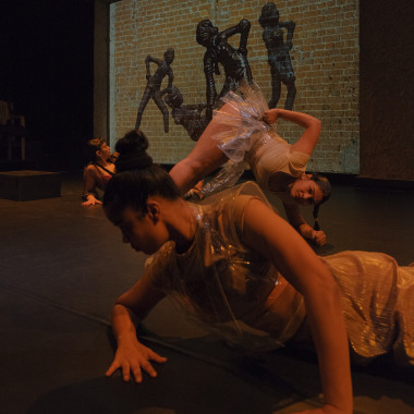 A stage situation: three dancers from the piece Hallucinations of an Artifact, behind them a brick wall on which the sculpture Dancing Girl is projected several times side by side. The performer in the foreground has tied her dark hair into a bun and is lying sideways on the floor. She is leaning on the floor with her hands spread wide apart and looking down. The person in the middle is leaning on the floor with her feet and hands, with the middle part of her body in the air. The third person in the background lies sideways on the floor and looks at the projection behind them. As with the projection of the dancing girl, one hand rests on the hip.