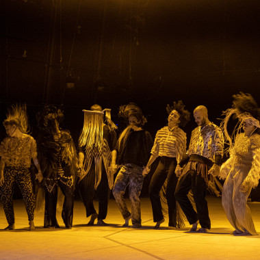 Eine Gruppe von sieben Performer*innen aus dem Stück TARAB auf einer Bühne, die in gelbes Licht eingetaucht ist. Sie stehen in einer Reihe und halten sich dabei an den Händen. Sie tragen weite Kleidung in Erdtönen und bewegen sich gleichzeitig. Dabei werfen sie ihre Haare in die Luft. 