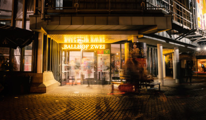 Das Ballhof Zwei-Gebäude bei Nacht. Über der Eingangstür aus Glas die gelbe, hell erleuchtete Aufschrift “Ballhof Zwei”.