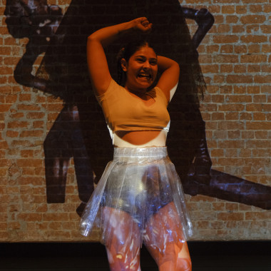 A dancing performer from the piece “Hallucinations of an Artifact”. The person raises their arms in the air and laughs. The feet are pointing in different directions. The person has long dark hair that is tied up. The person is wearing a beige-colored crop top and silver shorts. The person is wearing a transparent miniskirt around the hips. Behind the person is a brick wall on which several images of a sculpture are projected. 