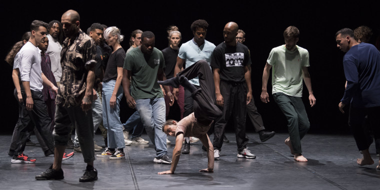 Eine Bühnensituation: Eine große Gruppe von Tänzer*innen aus dem Stück Apaches. Sie sind in weiten Hosen und lockeren Tshirts gekleidet. In der Bildmitte eine Person, die einen Handstand mit angewinkelten Beinen performt. Die Tänzer*innen um die Person herum beobachten ihn*sie und scheinen sich dabei zu bewegen.