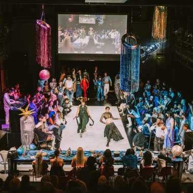 View of a catwalk situation from above. A long table is set up at the front of the picture, where the seven judges are sitting. In front of them is a white catwalk area. Two people in dresses and skirts are posing on it. Many other people are standing around them, clapping and cheering them on.