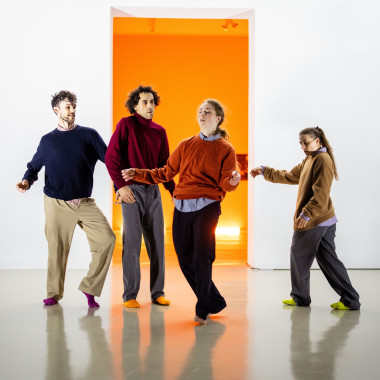 Die vier Performer*innen Yuri Fortini, Talal Mouzanar,Nina Melcher und Vivien Kaluza stehen in einer Reihe vor einer weißen Wand und tanzen jede*r für sich. Sie fragen bunte Socken, einfarbige Pullover in Blau, Rot, Orange und Braun. Im Hintergrund ist ein erleuchteter Raum mit orangefarbenen Wänden zu sehen.