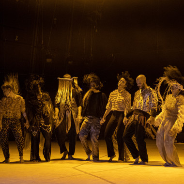 A group of seven performers from the play TARAB on a stage bathed in yellow light. They stand in a row, holding hands. They wear loose clothing in earth tones and move at the same time. They throw their hair in the air. 