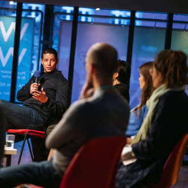 Podium situation: Tiago Manquinho speaks into a microphone and looks into the panel. The other panellists can only be seen from behind.